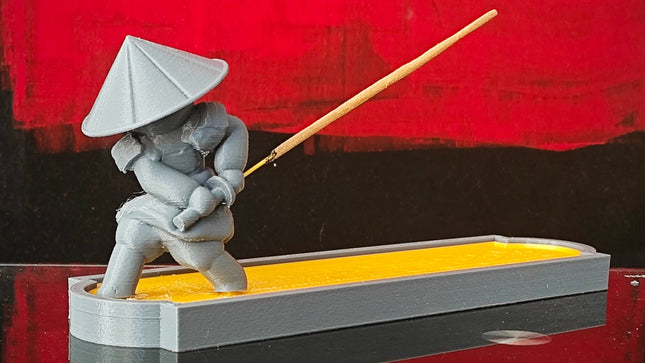 Sculpture of a person in traditional attire with an incense stick on a reflective surface against a red background